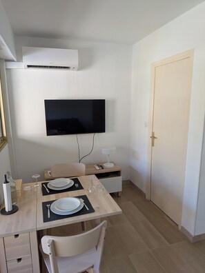 A dining area with a flat-screen TV mounted on the wall, a table set with plates, glasses, and cutlery, and a chair. - Charming 1-bedroom apartment with AC in peaceful Menton Garavan. (Menton)