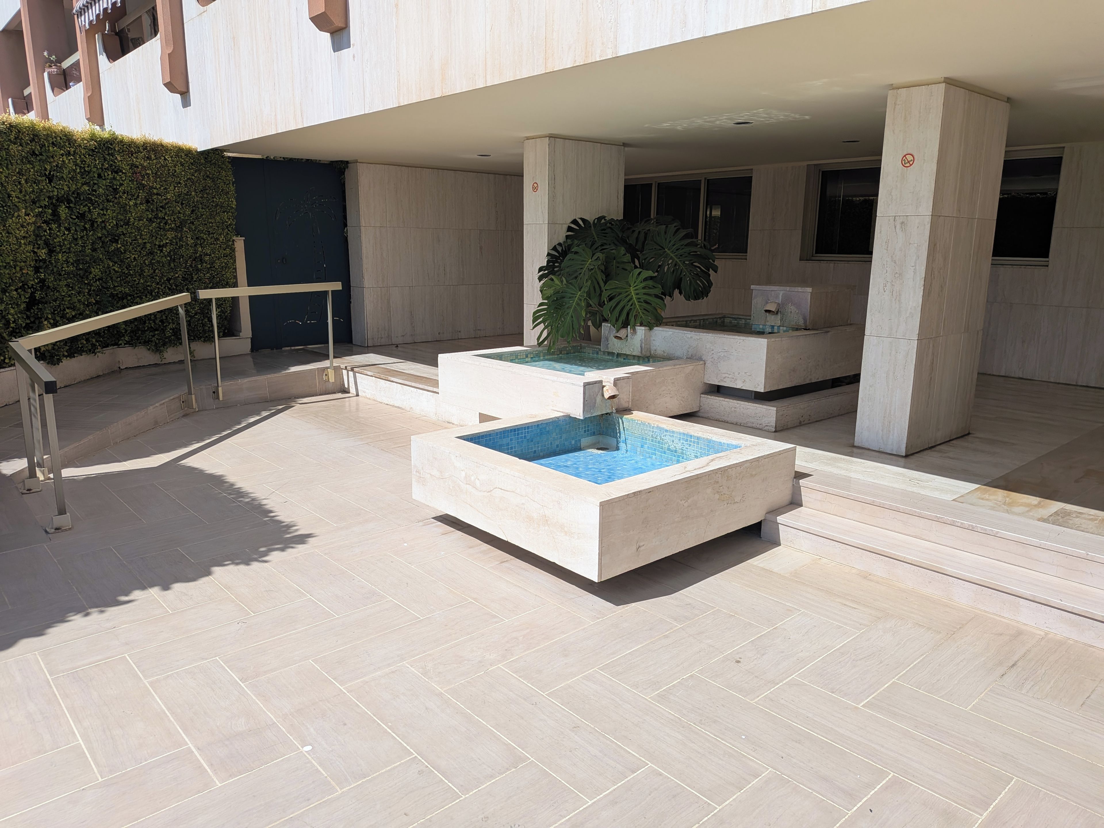 A modern outdoor pool area with a geometric design, surrounded by a low hedge and a metal railing.