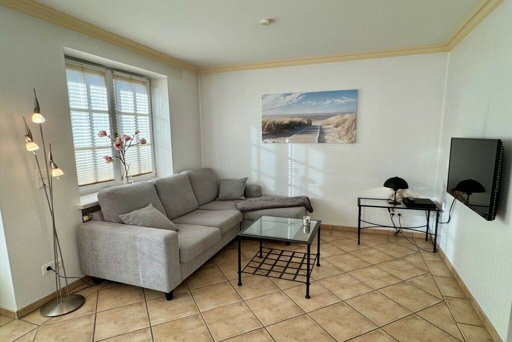 Un salon avec un canapé d’angle gris, une table basse en verre, un lampadaire, un tableau accroché au mur et un téléviseur posé sur un meuble.