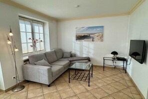 Una sala de estar con un sofá seccional gris, una mesa de centro de vidrio, una lámpara de pie, un cuadro en la pared y un televisor sobre un mueble.