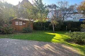 A small wooden cabin with a brick pathway leading to it, surrounded by greenery and a hedge. - Hansen Friesen House (Westerland)