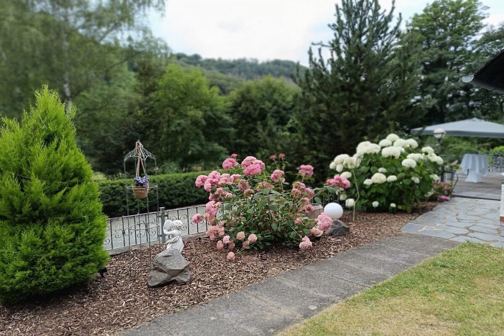 Un jardín con una variedad de plantas, incluyendo un arbusto grande con flores rosadas, un árbol de coníferas y un matorral con flores blancas.