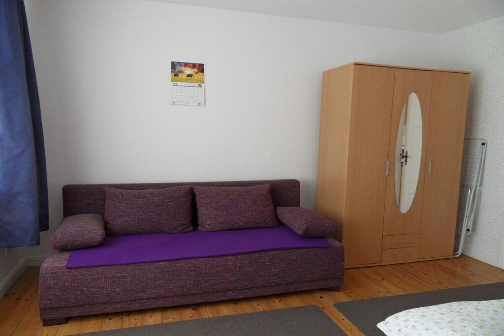 Une pièce avec un canapé violet, une armoire en bois, un calendrier accroché au mur et une fenêtre avec un rideau bleu.