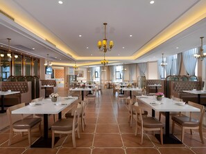 A spacious restaurant with multiple dining tables and chairs, a central chandelier, and large windows allowing natural light.
