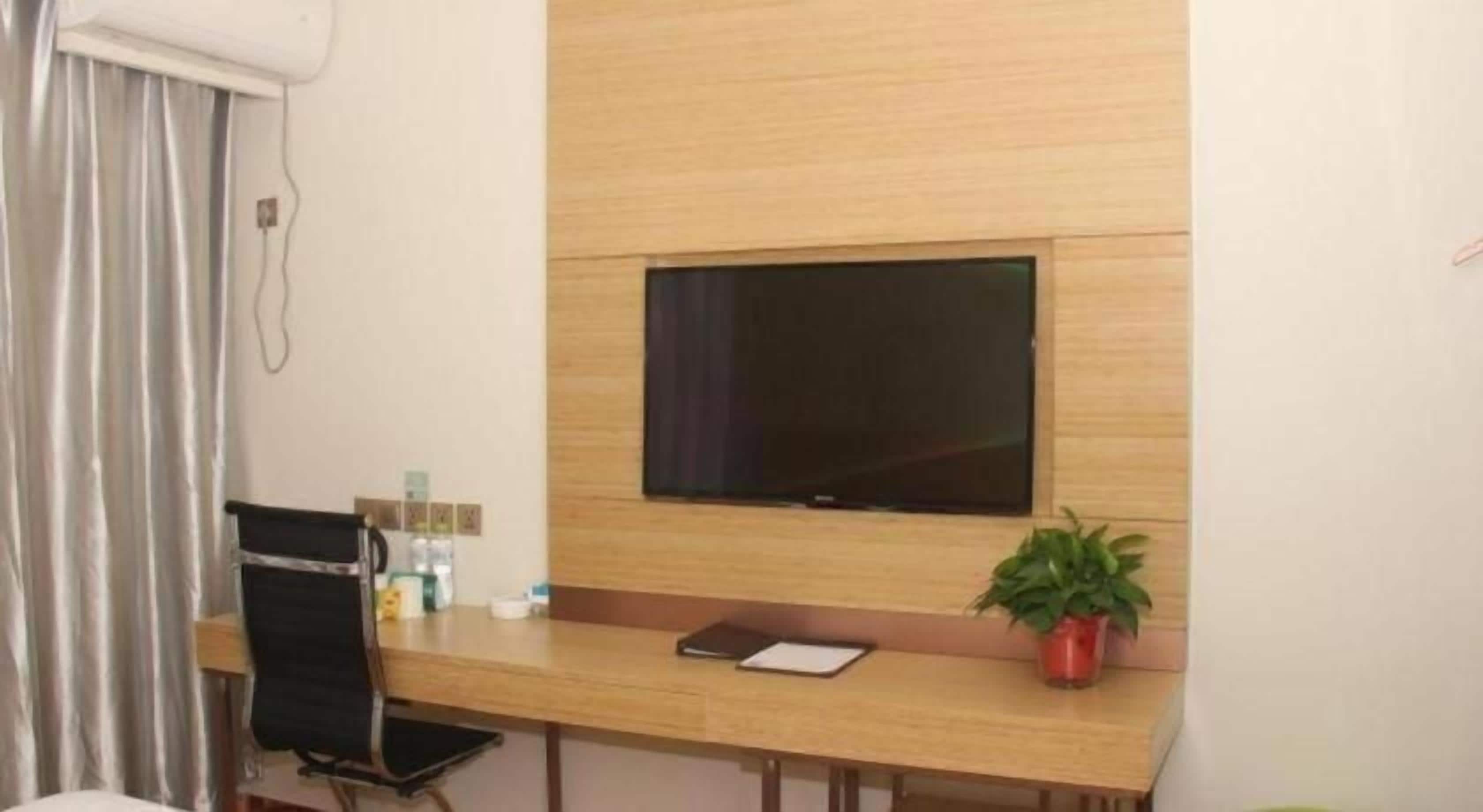A hotel room with a wooden desk, a television mounted on the wall, a chair, a potted plant, and a window with curtains.