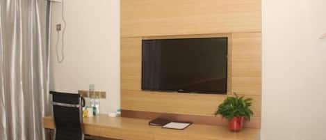 A hotel room with a wooden desk, a television mounted on the wall, a chair, a potted plant, and a window with curtains.