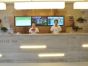A reception desk with two staff members, a digital screen displaying sports content, and a sign indicating the hotel name.