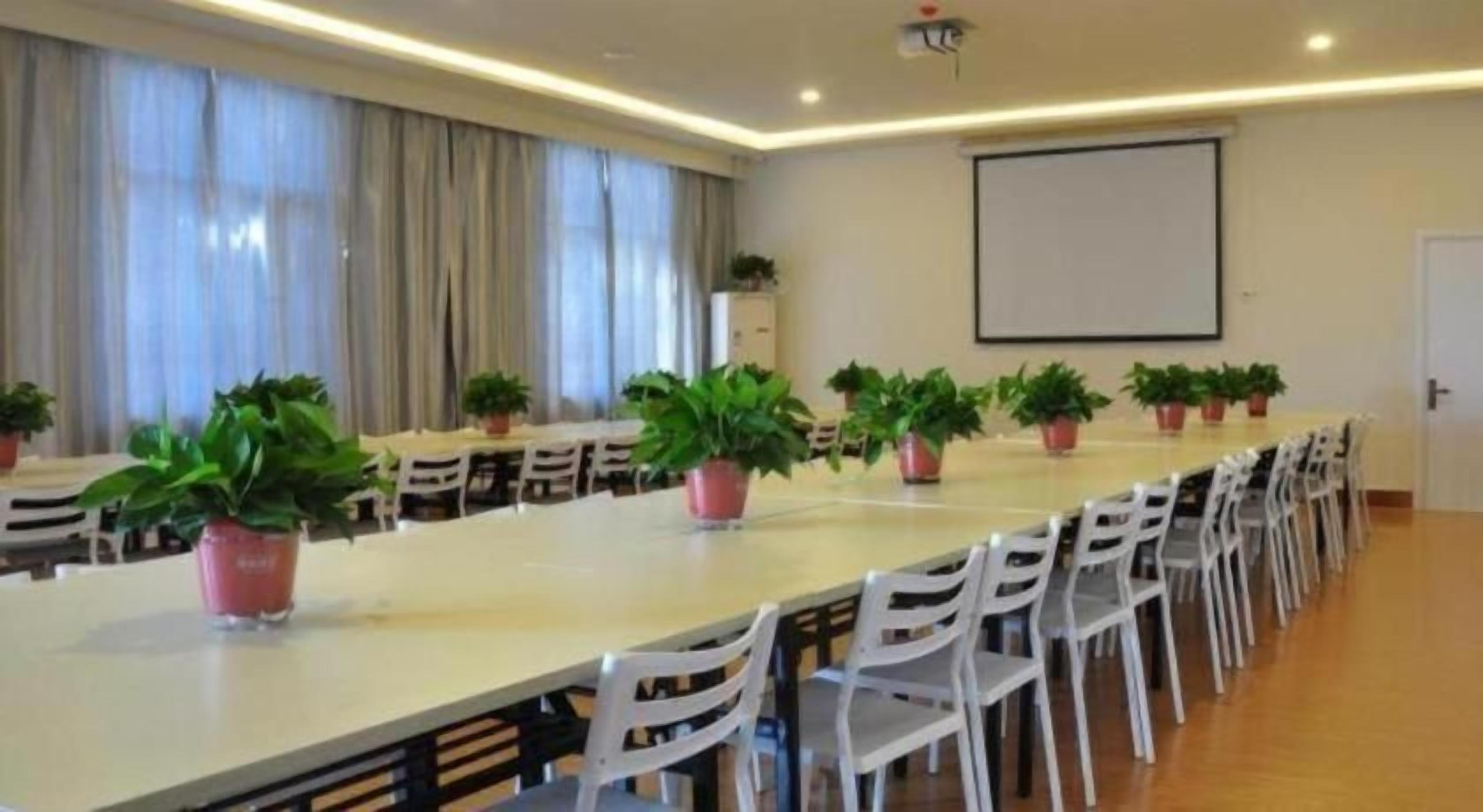 A conference room with a long table, white chairs, potted plants, a projector screen, and a large window with curtains.