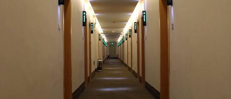 Un pasillo de hotel con puertas señalizadas con luces, pisos alfombrados y un camino recto hacia una salida distante.