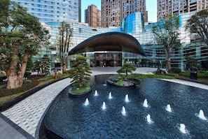 A modern building with a glass facade, a water feature with fountains, and a landscaped area with trees.