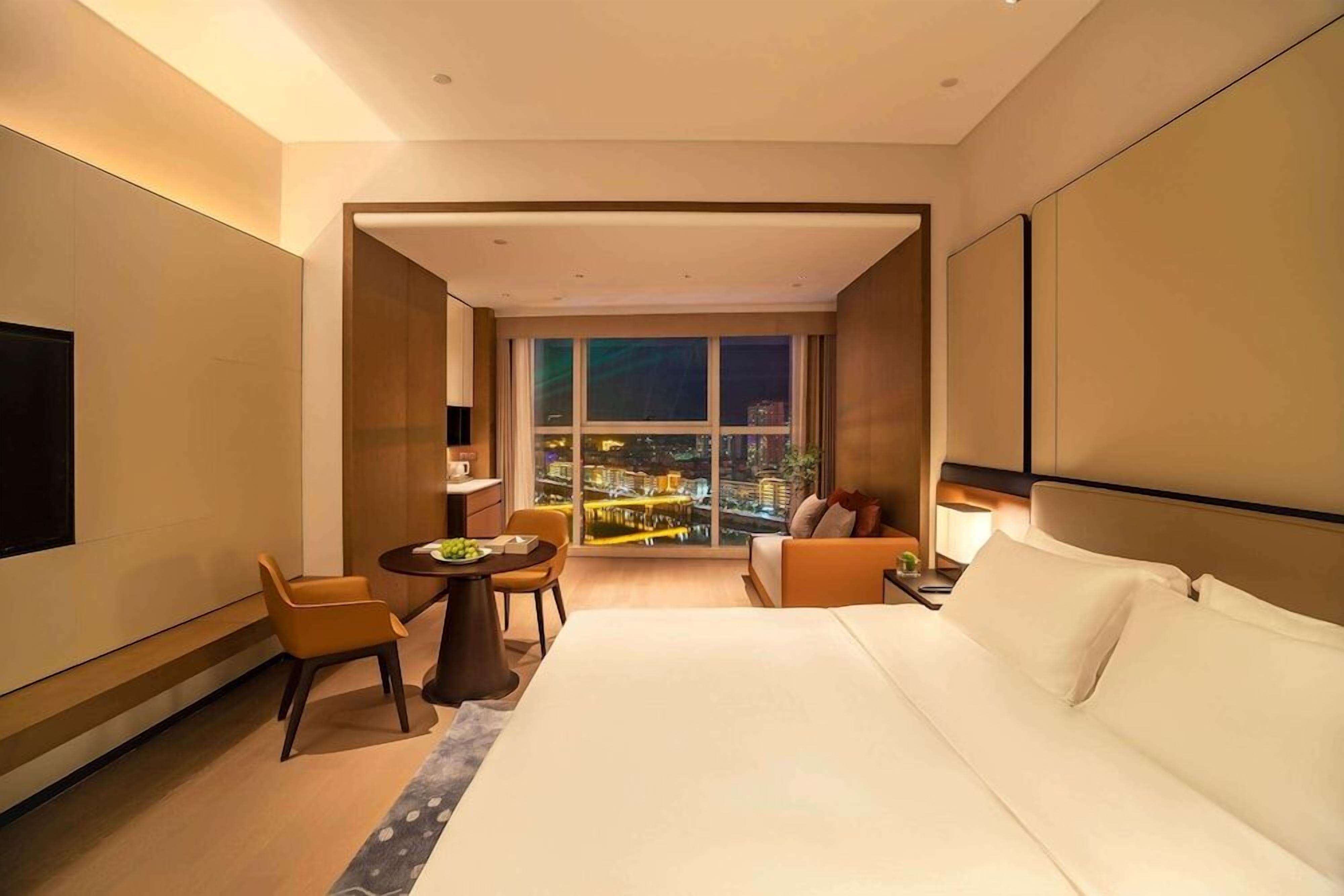 A hotel room with a large bed, a seating area with a table, and a view of a cityscape at night.