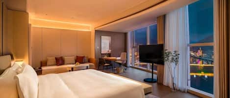 Quarto de hotel moderno com uma cama grande, escrivaninha, TV de tela plana e vista para a paisagem urbana à noite.