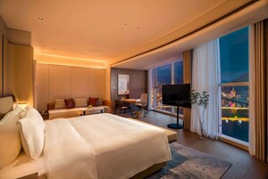 A modern hotel room with a large bed, a desk, a flat-screen TV, and a view of a cityscape at night.