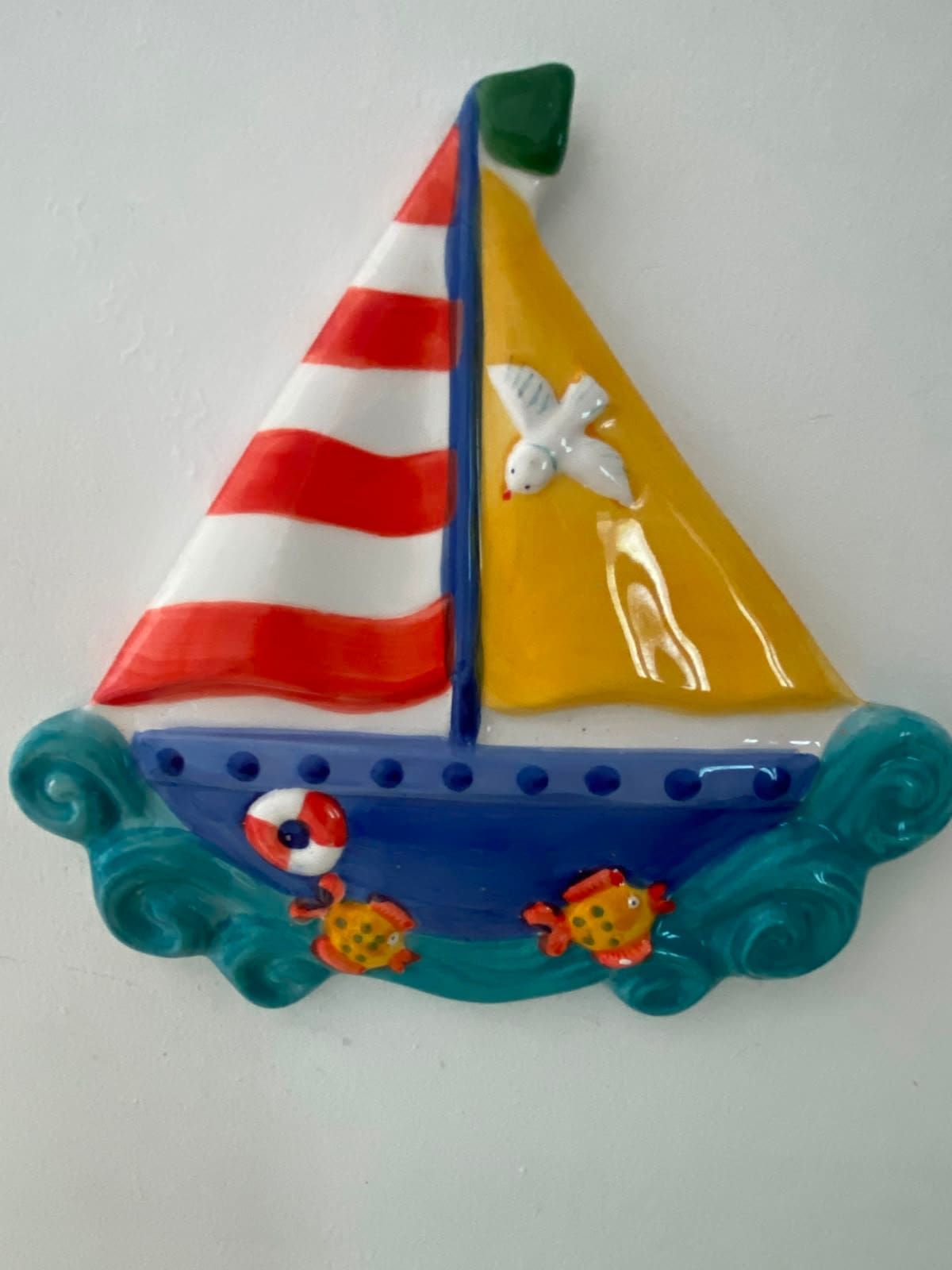 Ein dekoratives Segelboot mit einem gestreiften Segel, einem weißen Vogel und zwei Fischen.