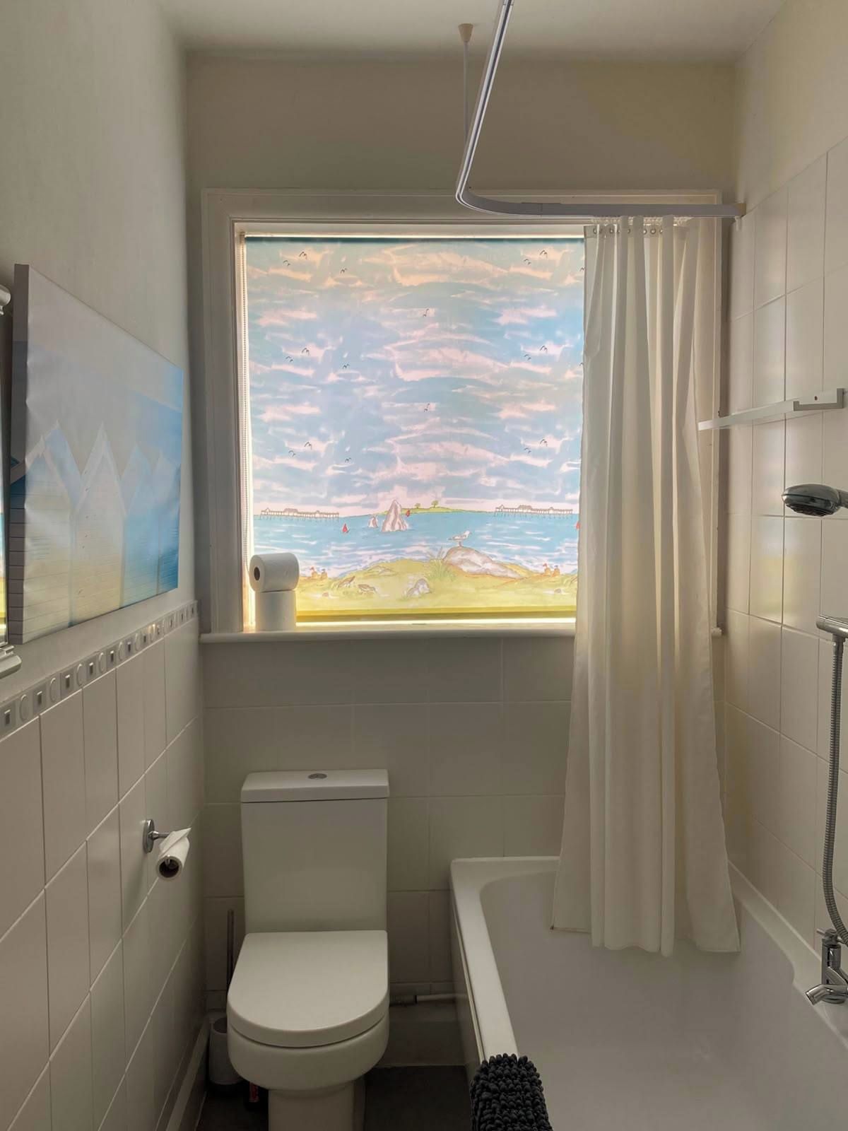 Ein Badezimmer mit Toilette, einer Badewanne und einer Dusche. Ein Fenster bietet Blick auf einen Strand und einen Steg.
