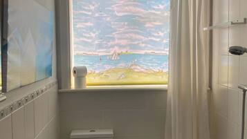 Ein Badezimmer mit Toilette, einer Badewanne und einer Dusche. Ein Fenster bietet Blick auf einen Strand und einen Steg.