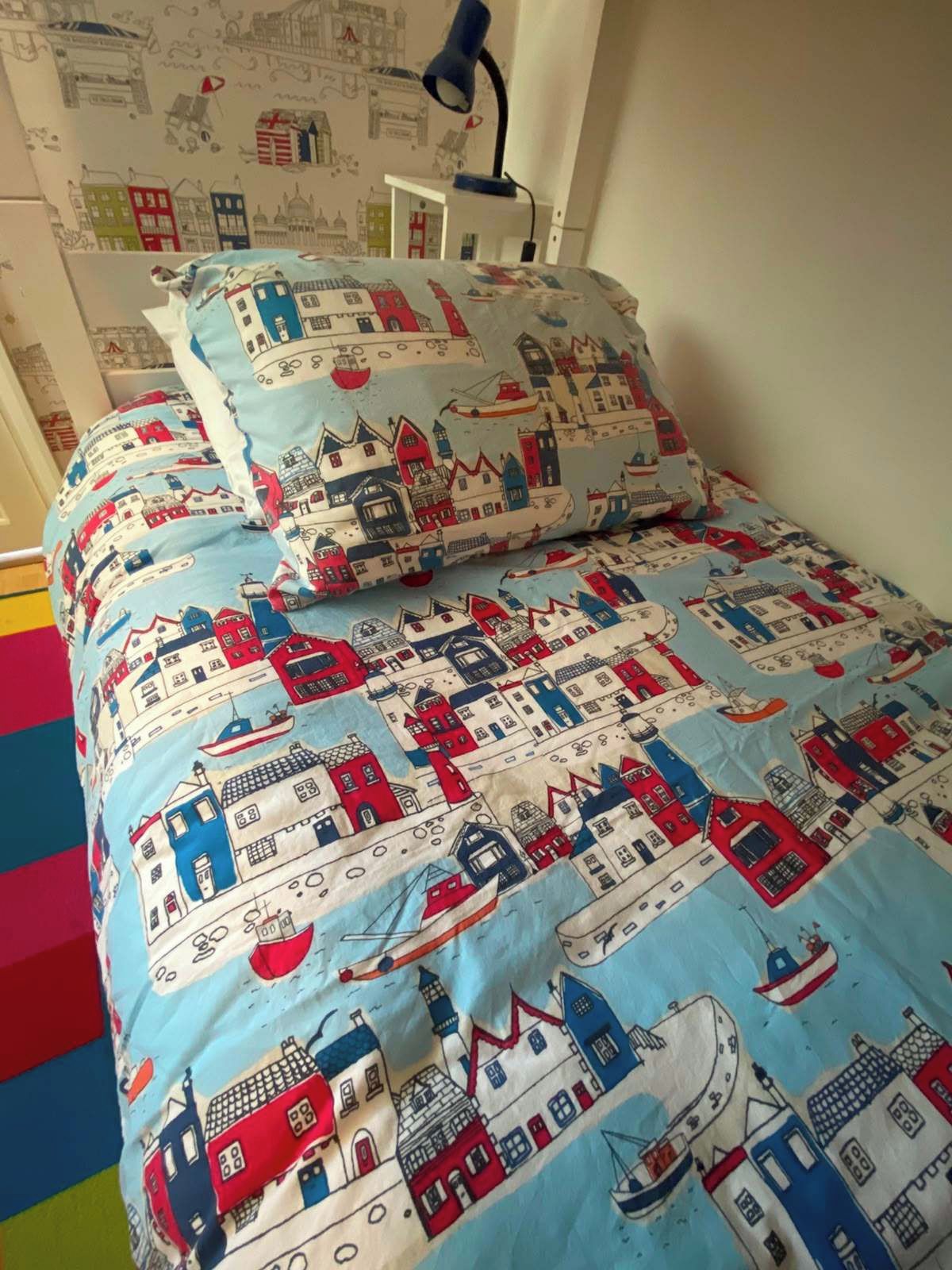 Un lit avec une couette et des oreillers à motifs bleus et blancs, une lampe de bureau et un mur recouvert d’un papier peint représentant un paysage urbain.