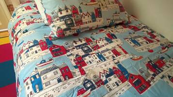 Un lit avec une couette et des oreillers à motifs bleus et blancs, une lampe de bureau et un mur recouvert d’un papier peint représentant un paysage urbain.