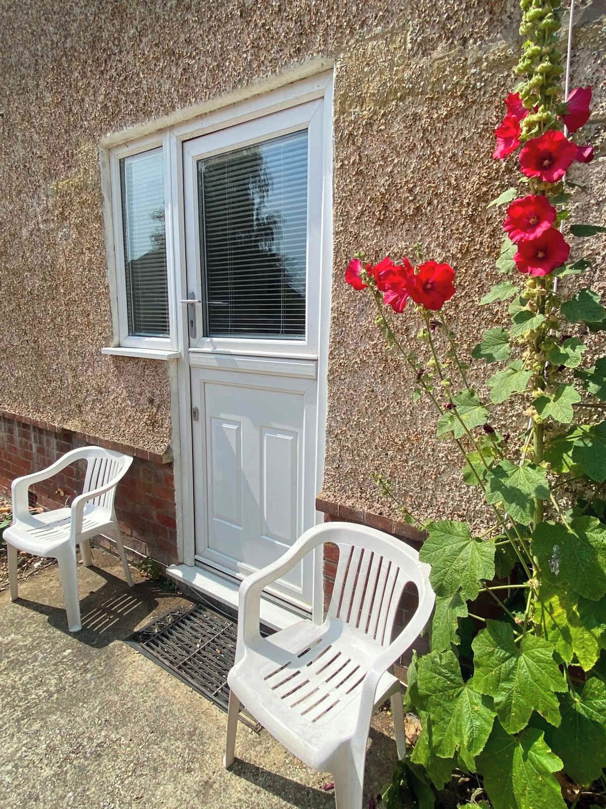 A white door with a window, two white plastic chairs, and a plant with red flowers.