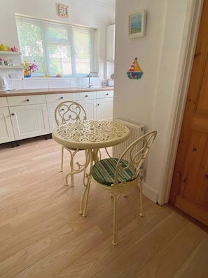 A dining area with a round table and chairs, a kitchen in the background, and a wooden door. - Spacious coastal apartment with garden, sleeps 6 (West Sussex)