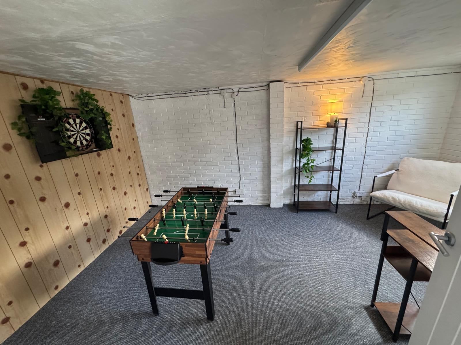 Una habitación con mesa de futbolín, sofá, estantería con plantas y dardo.