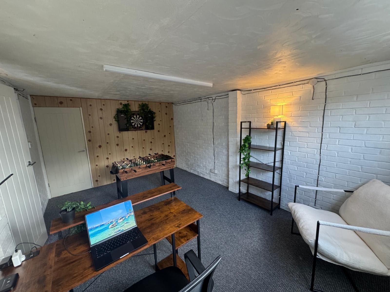 Una habitación con mesa de fútbol, escritorio con laptop, silla y estantería con plantas.