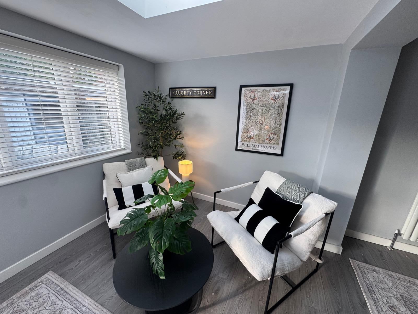 A room with a grey wall, a window with blinds, a black round table, two white armchairs with black cushions, a potted plant, a framed picture, and a sign that reads 'NAUGHTY CORNER'.