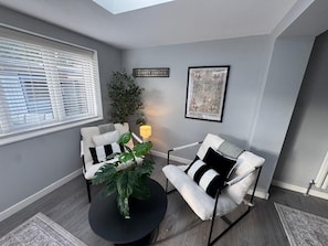 A room with a grey wall, a window with blinds, a black round table, two white armchairs with black cushions, a potted plant, a framed picture, and a sign that reads 'NAUGHTY CORNER'. - Lovely 5 bedroom holiday home with a games room (Christchurch)