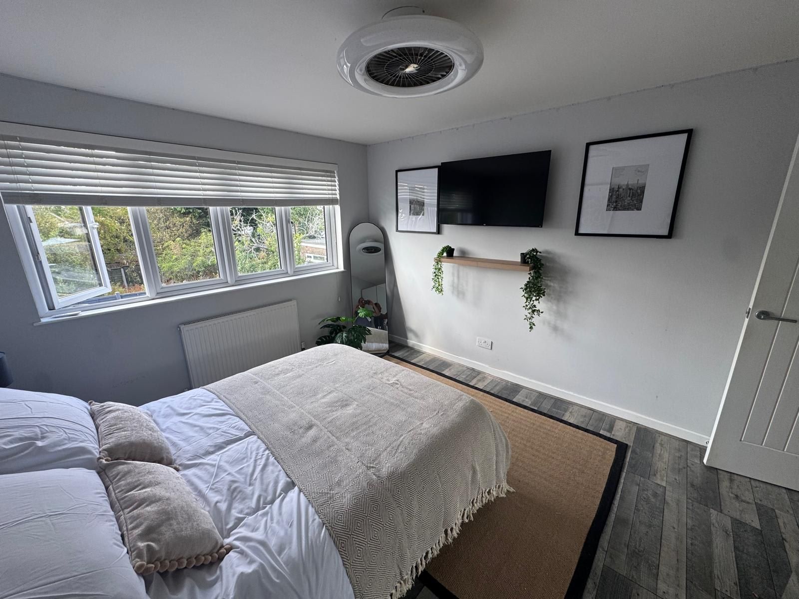 A bedroom with a bed, a radiator, a window with blinds, a wall-mounted TV, and a shelf with plants.