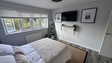A bedroom with a bed, a radiator, a window with blinds, a wall-mounted TV, and a shelf with plants.