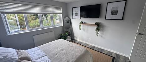 A bedroom with a bed, a radiator, a window with blinds, a wall-mounted TV, and a shelf with plants.