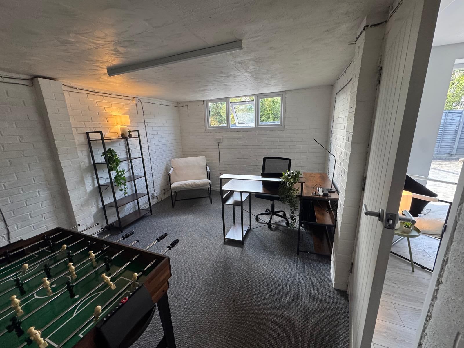 A room with a foosball table, a desk with a chair, a bookshelf, and a window.