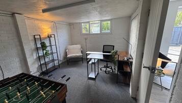 A room with a foosball table, a desk with a chair, a bookshelf, and a window.