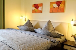Ein Bett mit grauer, gemusterter Bettdecke und Kissen, einem Kopfteil und zwei an der Wand befestigten Blumenbildern.