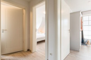 Ein Flur mit weißen Türen, der zu einem Schlafzimmer mit Bett und Fenster führt.