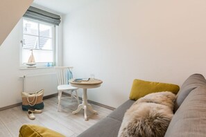 Ein gemütliches Zimmer mit einem grauen Sofa, einem kleinen runden Tisch, einem Sessel und einem Fenster mit Blick auf ein Gebäude.