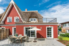 Ein rotes Haus mit einem Strohdach, einem Balkon und einer Terrasse mit Tisch und Stühlen.