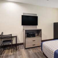Habitación de hotel con televisor de pantalla plana montado en la pared, microondas en un mueble, escritorio con silla y cama con colcha estampada.