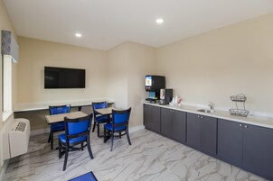 Habitación de hotel con comedor, televisor de pantalla plana en la pared, cafetera y una pequeña encimera con lavabo.