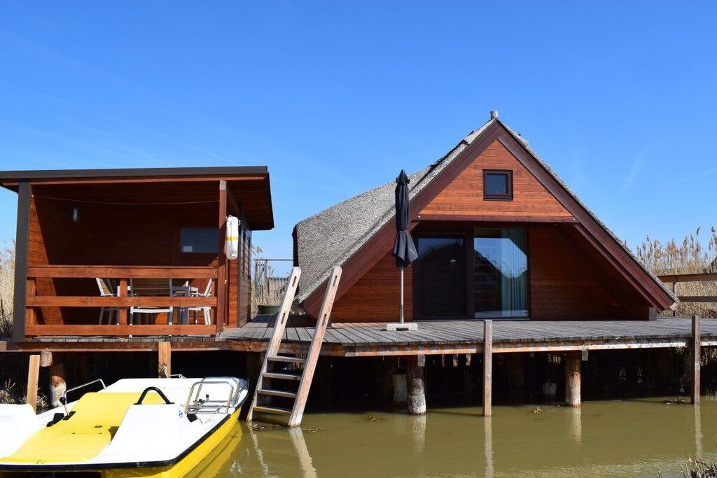 A wooden cabin with a deck, a boat docked in front, and a ladder leading to the cabin.