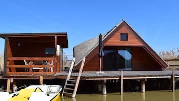 A wooden cabin with a deck, a boat docked in front, and a ladder leading to the cabin.