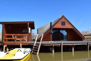 A wooden cabin with a deck, a boat docked in front, and a ladder leading to the cabin.
