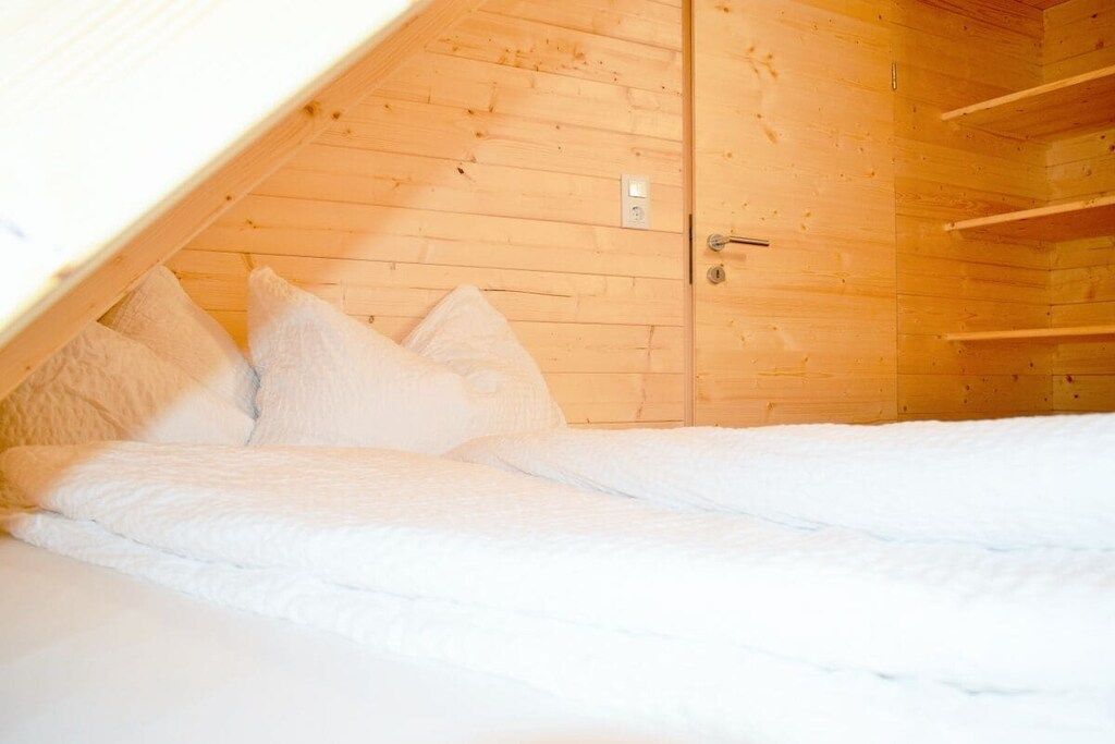 Une chambre avec un plafond en pente, des murs en bois, un lit avec une literie blanche et une porte dotée d’une poignée.