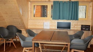 A wooden cabin with a dining area featuring a table and chairs, a television, and a window with curtains.