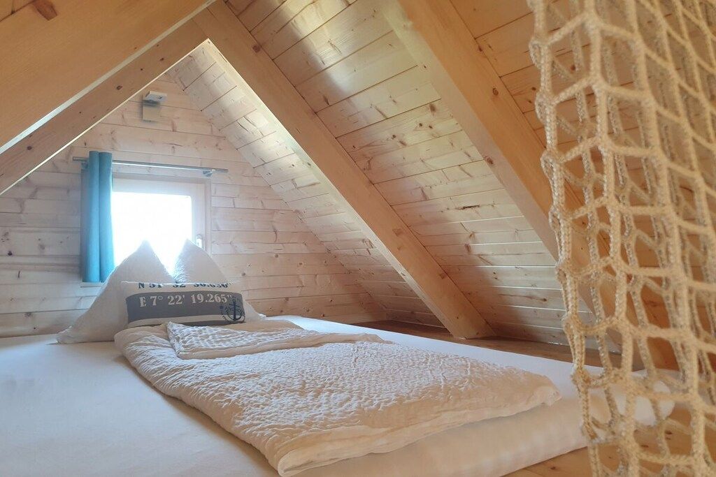 Ein gemütliches Schlafzimmer im Dachgeschoss mit schrägen Wänden, einem Bett mit weißer Decke und einem Fenster mit blauen Vorhängen.