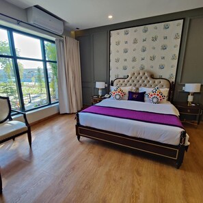 A bedroom with a large bed, a window with curtains, a chair, and a nightstand with a lamp.