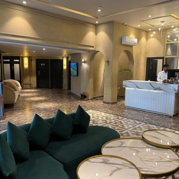 Un hall de hotel con mostrador de recepción, un sofá verde, suelo de mármol y un espejo grande.