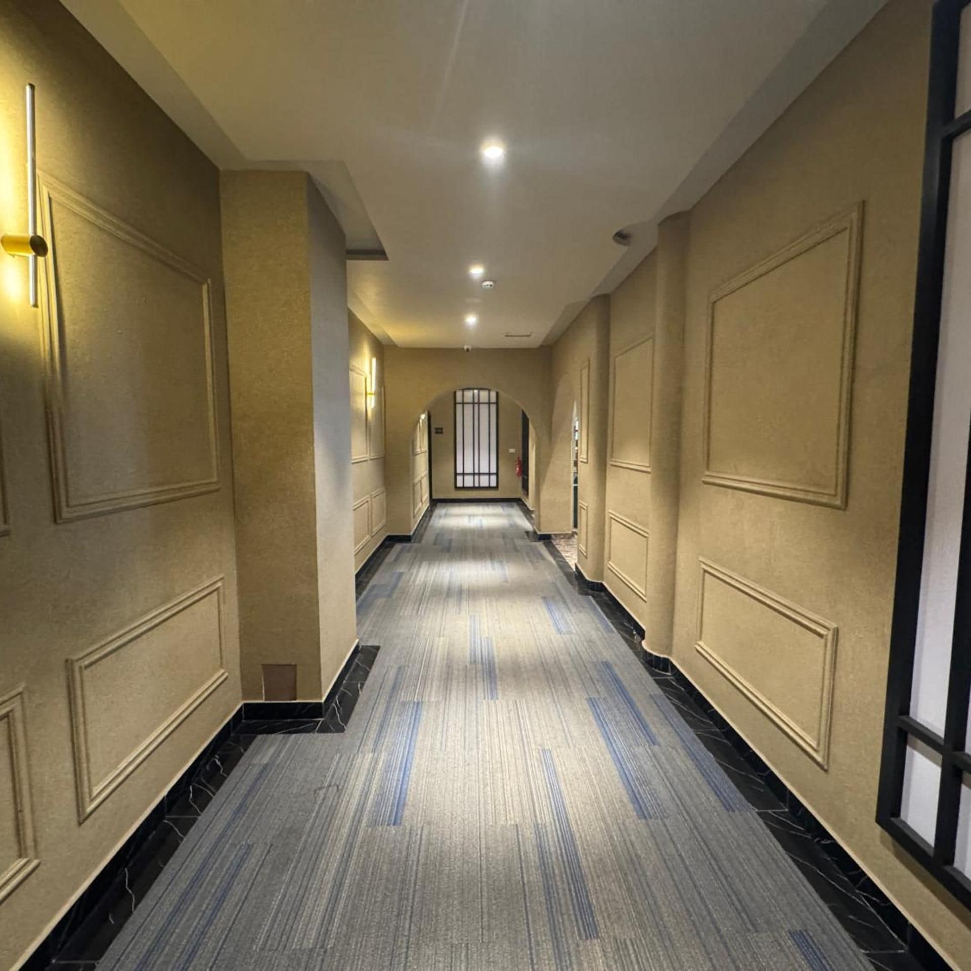 Ein Hotelgang mit Teppichboden, Wandleuchten und einer Tür am Ende.