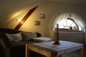 Ein gemütliches Zimmer mit schrägen Wänden, einem Holztisch, einer Couch mit Kissen, einer Lampe, einem Fenster mit Vorhängen und einer Heizung.