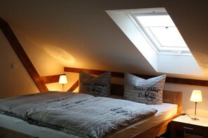 Ein Schlafzimmer mit schrägem Dach, einem Oberlicht, einem Bett mit gemustertem Überwurf, zwei Nachttischlampen und Kissen mit Schrift.